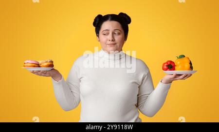 Une fille obèse choisissant entre deux assiettes avec des légumes frais et des beignets Banque D'Images