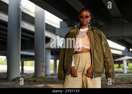 Femme afro-américaine portant une tenue décontractée debout sous un pont dans un cadre urbain montrant un comportement détendu et confiant Banque D'Images