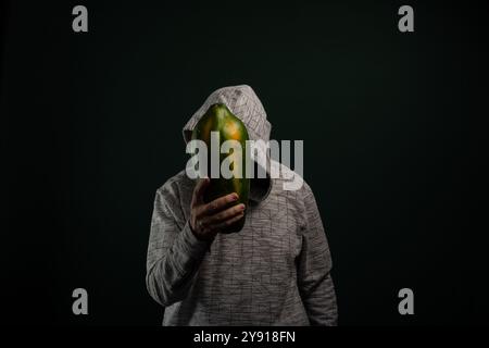 Homme mystérieux avec des lunettes portant un sweat à capuche tenant la papaye verte devant le visage. Isolé sur fond sombre. Banque D'Images