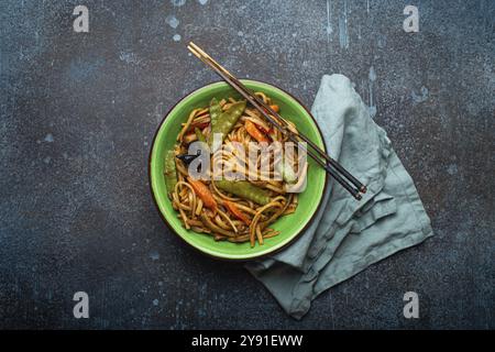 Plat asiatique sautés faire frire les nouilles udon avec poulet, légumes et champignons dans un bol en céramique verte avec des baguettes en bois sur fond en béton bleu foncé rustique Banque D'Images