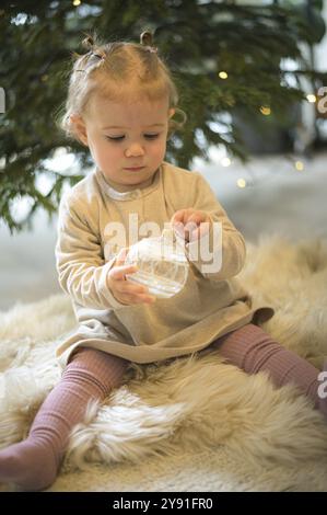 Un tout-petit tient une boule de Noël scintillante devant un sapin de Noël dans une atmosphère hivernale et chaleureuse Banque D'Images