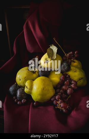 Une nature morte riche avec des coings fraîchement cueillis, des figues et des raisins disposés sur un tissu Bordeaux, capturés dans un éclairage chiaroscuro discret, avec drame Banque D'Images