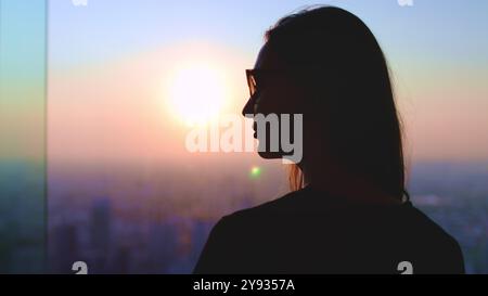 Femme profiter de la ligne d'horizon aérienne de la ville de coucher de soleil debout près de la fenêtre. Silhouette de fille à cheveux longs dans des lunettes de soleil contre le ciel coloré lumineux, le soleil se couche. Banque D'Images