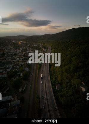 Vue aérienne d'une autoroute qui courbe à travers une forêt verdoyante et une ville au coucher du soleil, mettant en évidence des paysages urbains et naturels contrastés. Banque D'Images