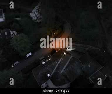 Vue aérienne de nuit d'une route de banlieue sombre et sinueuse avec un lampadaire chaud éclairant une section, entourée d'arbres et de toits résidentiels. Banque D'Images