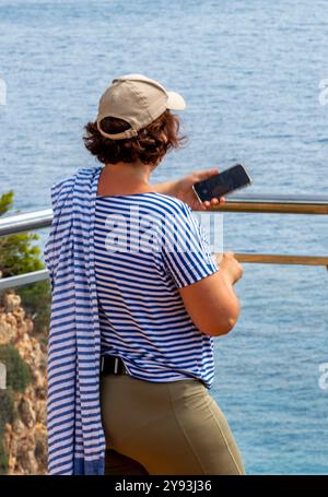 Femme d'âge moyen portant une chemise rayée bleue et blanche et une casquette de baseball prenant une photo à l'aide d'un smartphone depuis une falaise en Grèce. Banque D'Images
