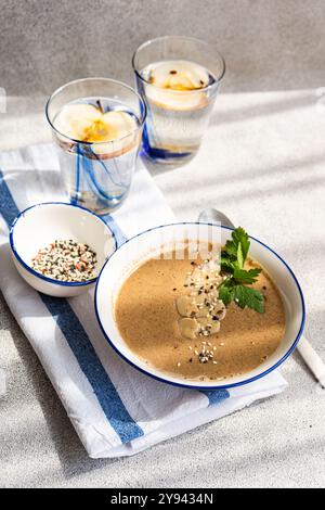 Un service de dîner sain comprenant une soupe crémeuse aux champignons garnie de graines et d'herbes dans un bol blanc, servi avec de l'eau de pomme dans des verres et un côté de Banque D'Images