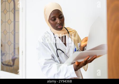 Une médecin noire concentrée portant un hijab examine les documents des patients dans un cadre clinique lumineux, incarnant le professionnalisme et les soins Banque D'Images