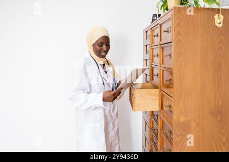 Une professionnelle médicale africaine en hijab et manteau blanc souriant alors qu'elle lit un document debout à côté d'un classeur en bois, elle représente Banque D'Images