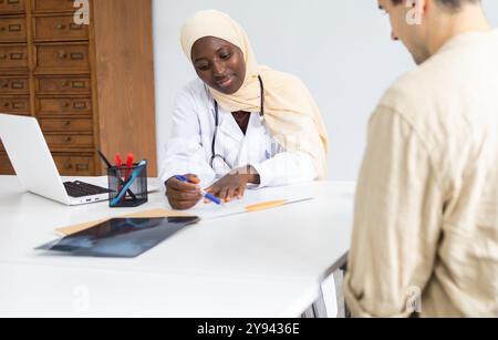 Une médecin noire professionnelle portant un hijab consulte attentivement un patient masculin dans son cabinet, expliquant les résultats obtenus à son pati Banque D'Images