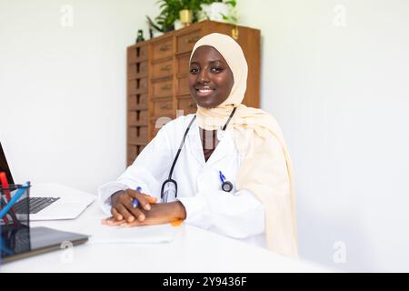 Une gaie médecin africaine portant un manteau blanc et le hijab est assise à un bureau dans une clinique lumineuse, préparée avec confiance pour aider les patients Banque D'Images