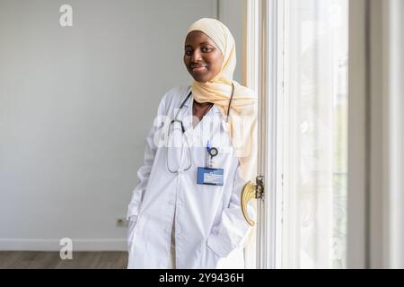 Une femme médecin confiante portant un hijab et un manteau blanc se tient près d'une fenêtre, représentant la diversité dans les soins de santé avec le sourire Banque D'Images