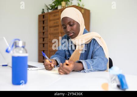 Une femme noire concentrée médecin portant un hijab est assise à son bureau, prenant diligemment des notes d'un livre elle est entourée d'objets, y compris un ordinateur portable et Banque D'Images