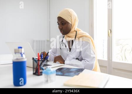 Médecin portant un hijab intensément concentré tout en travaillant sur son ordinateur portable dans un bureau lumineux et moderne Banque D'Images