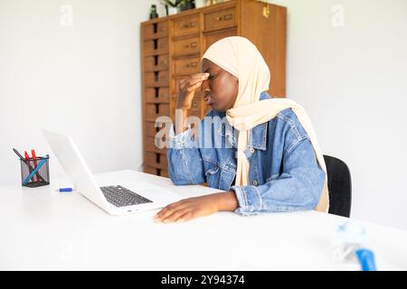 Médecin noire portant un hijab semble stressée tout en travaillant sur son ordinateur portable dans un bureau moderne elle est habillée dans une tenue décontractée en denim, illustratin Banque D'Images