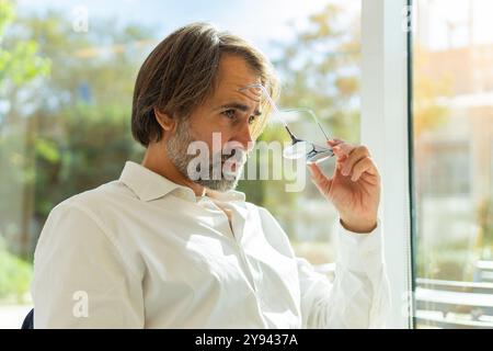 Homme d'affaires d'âge moyen regarde pensivement à travers une fenêtre dans un bureau lumineux, tenant ses lunettes dans une main. Banque D'Images