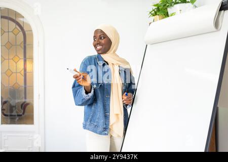 Une femme médecin noire souriante dans une veste en denim et des gestes hijab avec un marqueur à côté d'un tableau blanc dans une pièce lumineuse, suggérant une conférence engageante Banque D'Images