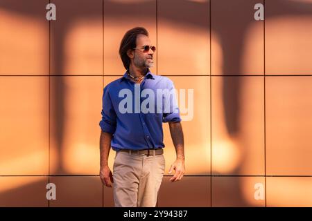 Un homme mûr, vêtu d'une chemise bleue décontractée et d'un pantalon beige, se promène en toute confiance dans une ville. Il porte des lunettes de soleil et une montre moderne, Banque D'Images