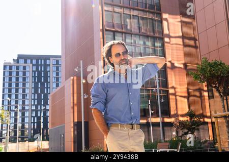 Un homme d'affaires mature avec une barbe et une chemise bleue décontractée apprécie la chaleur de la lumière du soleil dans un environnement urbain moderne, protégeant ses yeux et son regard Banque D'Images