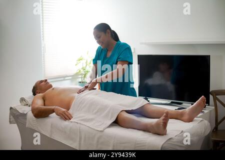 Un homme détendu allongé sur une table de massage obtenant un massage à la maison d'une thérapeute féminine souriante dans les gommages. L'homme a les yeux fermés et semble calme, Banque D'Images