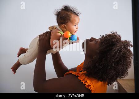 Une joyeuse mère africaine dans une blouse orange tient ludique sa petite fille en l'air de l'enfant, tenant des hochets colorés, regarde sa mère, créant une écoute Banque D'Images