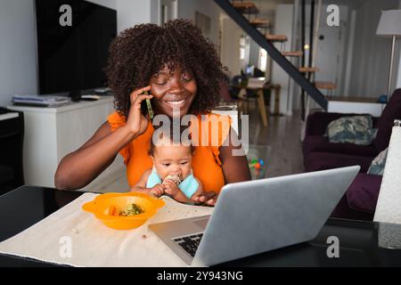 Une mère africaine noire joyeuse s'engage dans le multitâche, parlant au téléphone et regardant son bébé manger pendant qu'elle travaille sur un ordinateur portable dans un moderne, bien- Banque D'Images