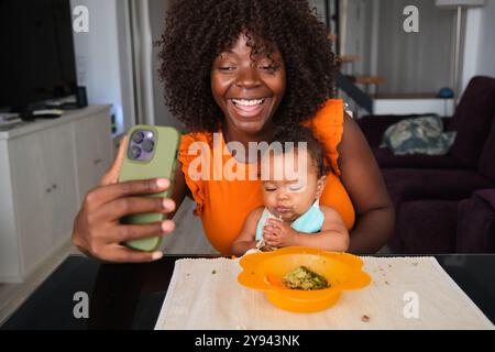 Une femme africaine prend joyeusement un selfie avec sa petite fille, qui mange dans un bol, capturant un moment joyeux et familial à la maison Banque D'Images