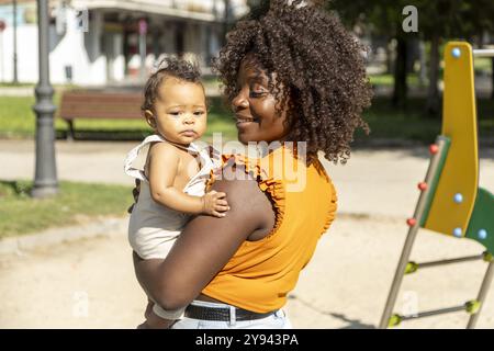 Mère africaine souriante dans un haut orange porte sa petite fille tout en profitant d'une journée ensoleillée dans le parc Banque D'Images