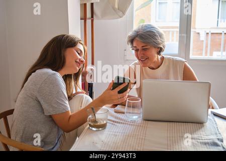 Une scène chaleureuse et engageante d’une mère et d’une fille à une table de cuisine, souriante et interagissant sur un smartphone, avec un ordinateur portable et des boissons présentes, co Banque D'Images