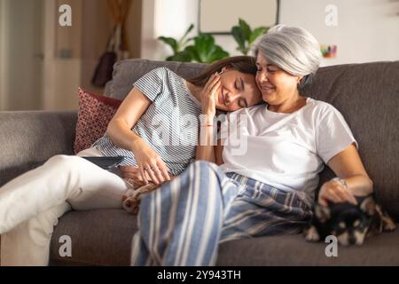 Une scène réconfortante d'une mère mature et de sa jeune fille adulte partageant un moment tendre sur un canapé, avec de petits chiens se reposant à côté d'eux Banque D'Images