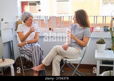 Une femme âgée souriante et sa jeune fille adulte discutent en toute décontraction tout en sirotant des boissons sur un balcon ensoleillé orné de plantes Banque D'Images