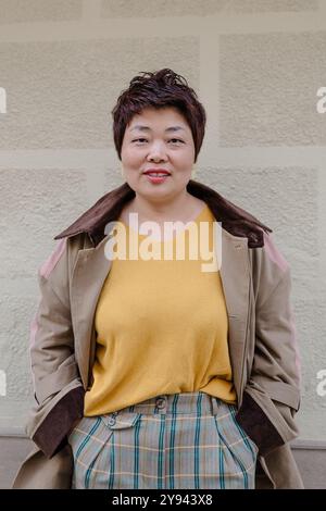 Une joyeuse femme asiatique d'âge moyen se tient contre un mur beige, vêtue d'un pull jaune et revêtue d'une élégante veste marron sur un plaid Banque D'Images
