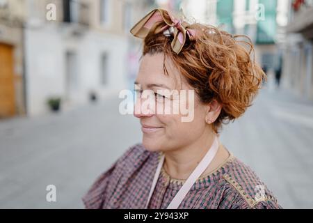 Un portrait franc d'une femme mature avec les cheveux roux bouclés et un ruban élégant, souriant doucement tout en se tenant debout à l'extérieur dans une rue de la ville Banque D'Images