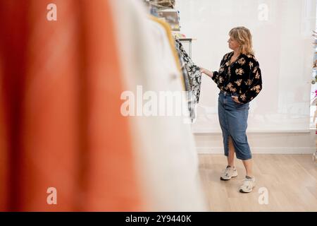 Une femme d'âge moyen habillée décontractée dans une jupe en Jean et un chemisier fleuri regardant les étagères à vêtements dans une boutique bien éclairée Banque D'Images