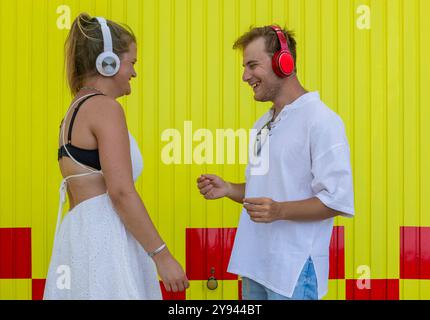 Un couple handicapé intellectuellement, tous deux portant des écouteurs, interagissent devant un mur jaune vif avec des bandes rouges la scène capture un an joyeux Banque D'Images