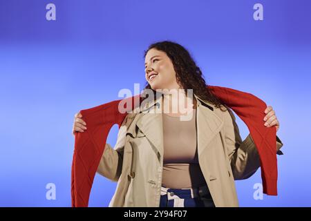 Une femme rayonnante de grande taille pose de manière ludique, tenant un pull rouge sur un fond éclatant. Banque D'Images