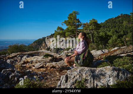 Une femme est assise paisiblement sur un terrain rocheux, en admirant le magnifique paysage montagneux autour d'elle. Le soleil brille, créant une atmosphère parfaite Banque D'Images