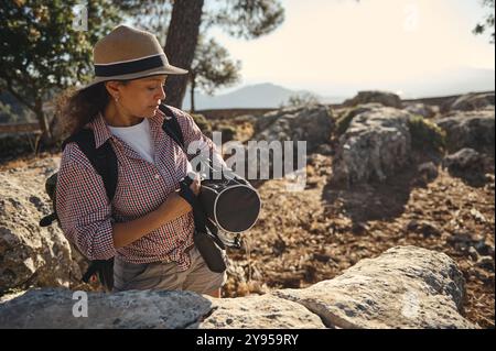 Une femme portant un chapeau explore un paysage rocheux, portant un sac à dos et un appareil photo. Elle aime la nature et l'aventure sous le ciel clair, capturant momen Banque D'Images