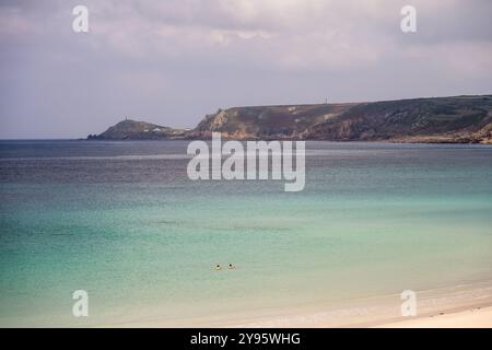 Les gens nagent dans la mer à Sennen Cove près de Land's End, avec le cap Cornwall promontoire au loin. Banque D'Images