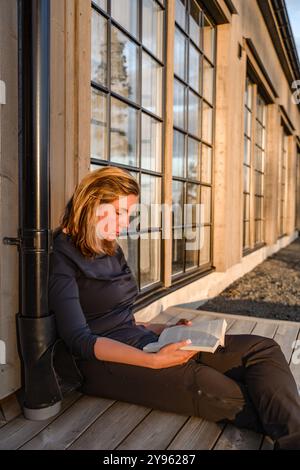 Belle femme est assise paisiblement sur une terrasse en bois, engrossée dans un livre que la lumière chaude du soleil couchant renforce l'atmosphère sereine autour d'elle Banque D'Images