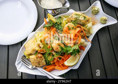 Grand plateau de poisson avec divers filets de poisson frits, légumes, citron, herbes et sauce, servi avec des assiettes blanches sur une table en bois foncé, vu de abo Banque D'Images