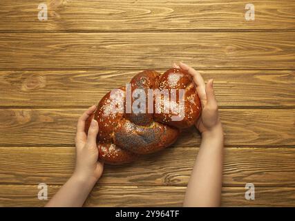 Mains féminines tenant du pain Challah fraîchement cuit recouvert de pavot et de graines de sésame, vue de dessus sur fond en bois rustique, juif traditionnel festif Banque D'Images