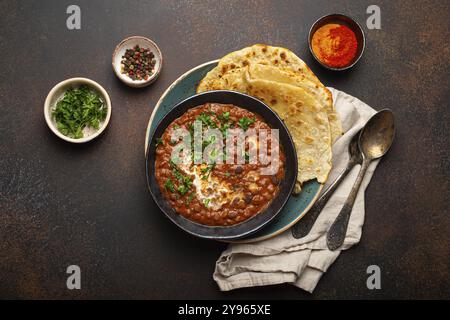 Plat traditionnel indien pendjabi Dal makhani avec lentilles et haricots dans un bol noir servi avec du pain plat naan, coriandre fraîche et deux cuillères sur c brun Banque D'Images