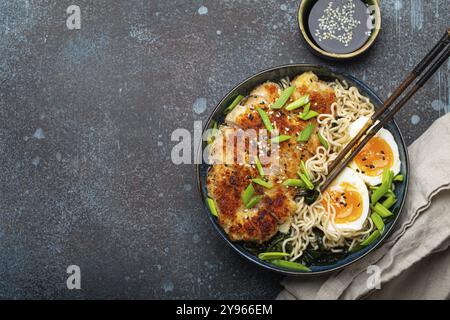 Soupe ramen aux nouilles asiatiques avec filet de poulet panko frit et œufs durs dans un bol en céramique avec des bâtons de côtelette et sauce soja sur fond rustique en pierre Banque D'Images