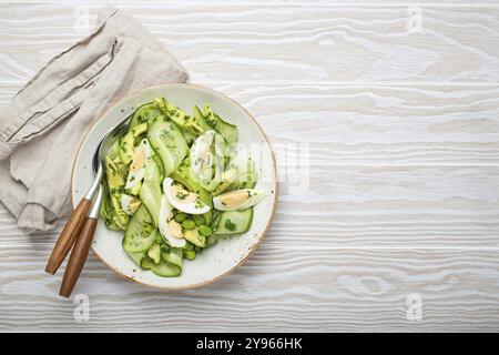 Bol à salade d'avocat vert sain avec œufs durs, concombres tranchés, haricots edamame, huile d'olive et herbes sur plaque en céramique vue de dessus sur la rouille en bois blanc Banque D'Images