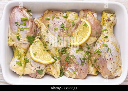 Cuisses de poulet crues non cuites marinées avec des assaisonnements, des herbes, du citron dans une casserole en céramique blanche vue de dessus sur fond rustique en bois clair. Préparation de HEA Banque D'Images