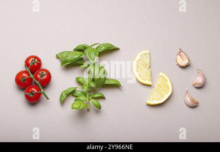 Composition avec des ingrédients alimentaires sains : branche de tomates cerises fraîches, branche de basilic, gousses d'ail, quartiers de citron sur dos gris minimaliste Banque D'Images