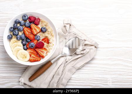 Porridge de flocons d'avoine avec fruits et baies dans un bol avec cuillère sur fond de bois blanc vue sur le dessus de la table, céréales de petit déjeuner saines maison avec paille Banque D'Images