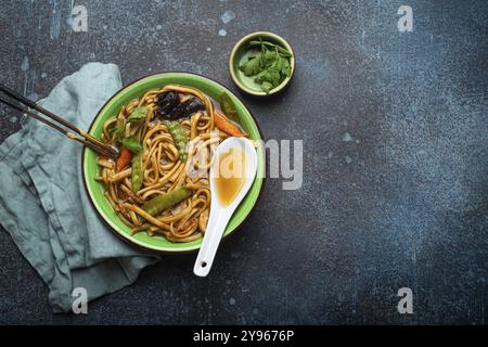 Soupe asiatique avec nouilles udon, poulet et légumes dans un bol en céramique verte avec baguettes en bois sur fond de béton bleu foncé rustique d'en haut c Banque D'Images