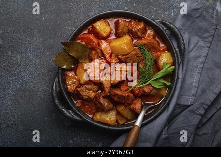 Ragoût de viande de bœuf avec pommes de terre, carottes et sauce délicieuse dans une casserole noire avec feuilles de Laurier et herbes vertes fraîches avec une cuillère sur noir rustique foncé Banque D'Images